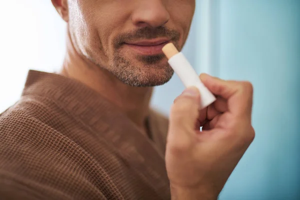 Hombre joven usando lápiz labial higiénico sobre fondo azul —  Fotos de Stock