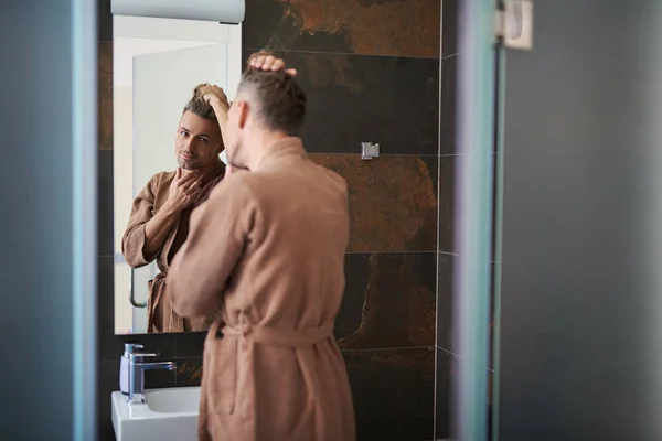 Guapo joven mirando en el espejo mientras está de pie cerca del lavabo del baño —  Fotos de Stock