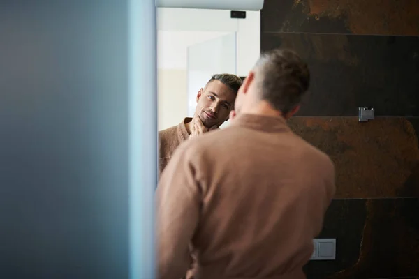 Hombre guapo sin afeitar mirando en el espejo y sonriendo un poco — Foto de Stock