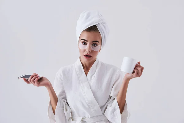 Jovencita desconcertada con parches bajo los ojos sosteniendo el teléfono celular y la taza de café — Foto de Stock