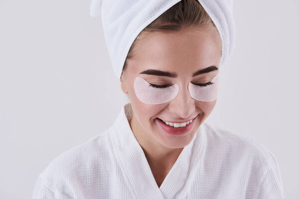 Joyful young lady with towel on head using under-eye patches