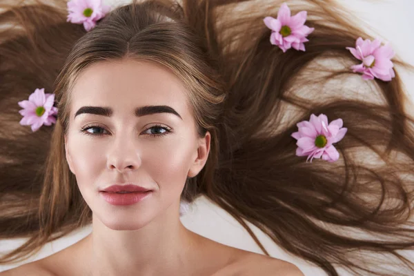 Charmante jonge dame met bloemen in haar haar liggend op witte achtergrond — Stockfoto