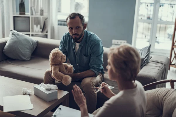 Hombre barbudo sosteniendo osito de peluche y escuchando a su psicoterapeuta — Foto de Stock