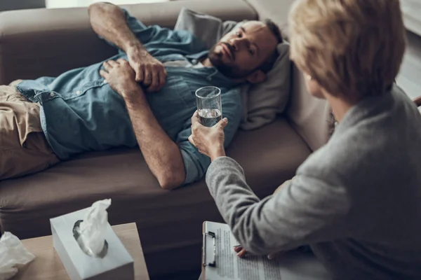 Hombre serio acostado en el sofá y mujer dándole un poco de agua — Foto de Stock