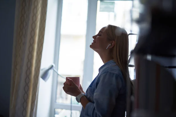 Beautiful blond girl with hot drink listening music through wireless earphones — Stock Photo, Image