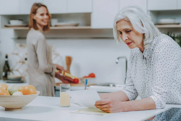 Пожилая дама пишет, пока ее дочь готовит в помещении — стоковое фото