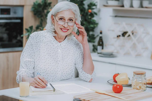 Vrolijke bejaarde vrouw is het schrijven van recept tijdens het koken — Stockfoto