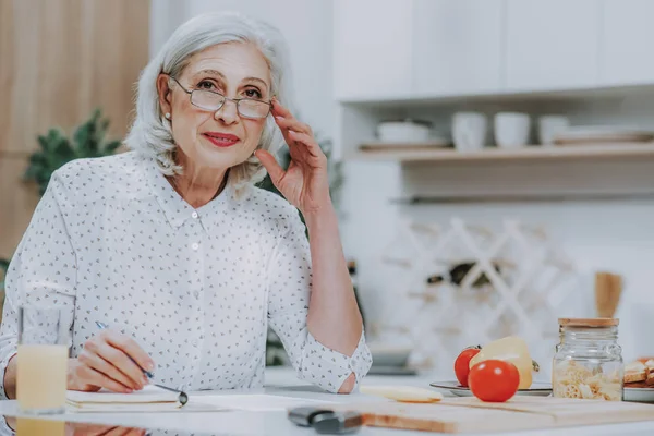 Mujer mayor sonriente está escribiendo en la mesa de la cocina — Foto de Stock