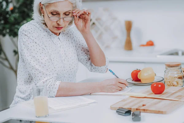 Aufmerksame ältere Frau schreibt beim Kochen in Skizzenbuch — Stockfoto