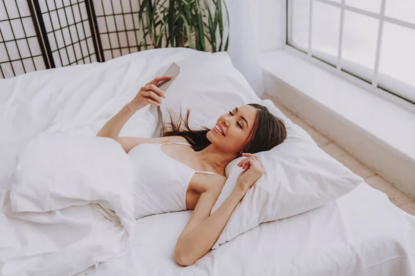 Улыбающаяся азиатка смотрит на мобильный телефон в спальне — стоковое фото