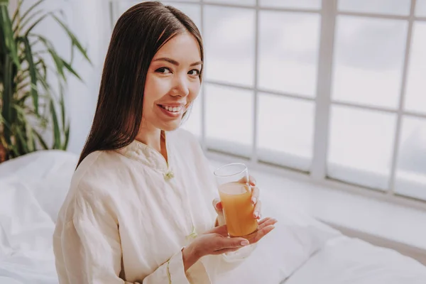 Cheerful young female holding glass of juice near window — Stock Photo, Image