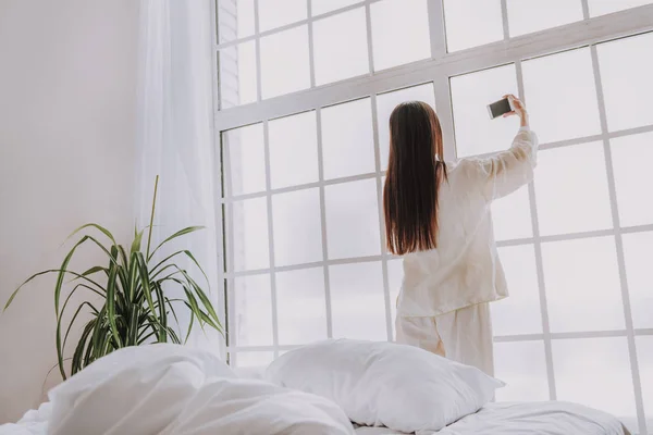 Calma mujer haciendo selfie por teléfono celular cerca de ventana ancha —  Fotos de Stock