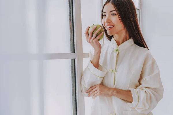 Щаслива молода дівчина дегустує яблуко біля широкого вікна — стокове фото