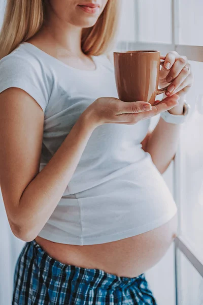 Беременная женщина держит чашку чая у большого окна — стоковое фото