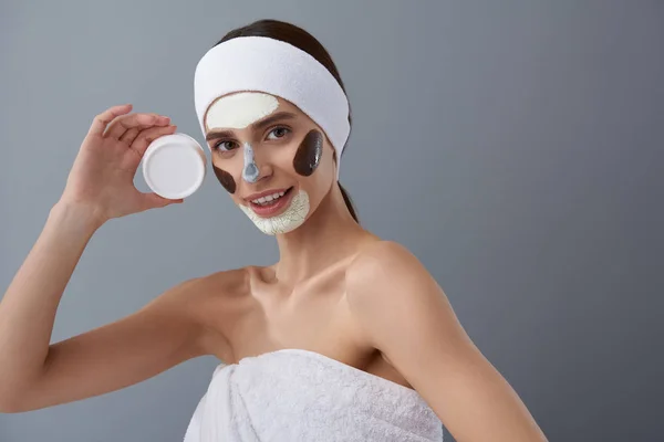 Joyful young lady with different masks on her face holding white jar of cream — Stock Photo, Image