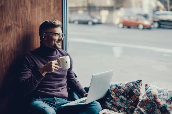 Homme assis dans un café avec ordinateur portable sur les genoux et tasse dans les mains — Photo