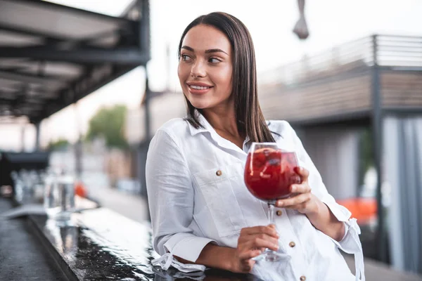 Waist up of relaxed young lady putting one elbow on the bar counter and holding her cocktail while smiling and looking away