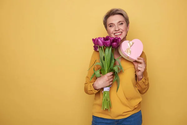 Waist up of smiling woman holding purple tulips and gift in arms