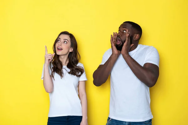 Casal interracial chocado em camisetas brancas posando para câmera em estúdio — Fotografia de Stock