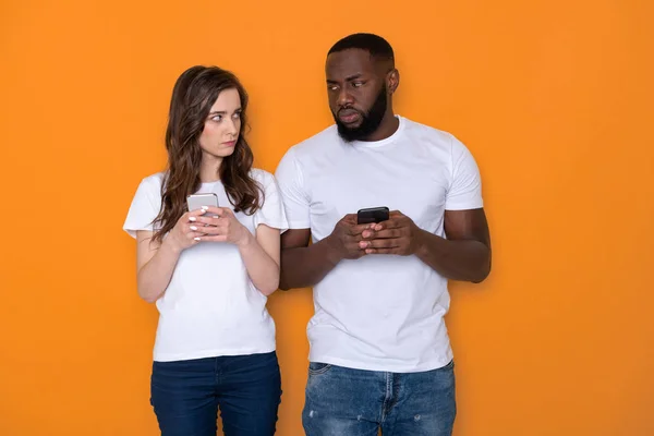 Couple interracial méfiant en T-shirts blancs tenant des smartphones dans les bras — Photo