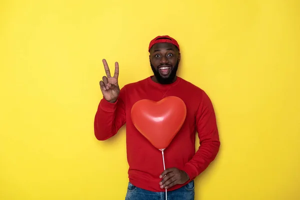 Sonriente hombre africano manteniendo globo rojo y mostrando señal de mano victoria — Foto de Stock