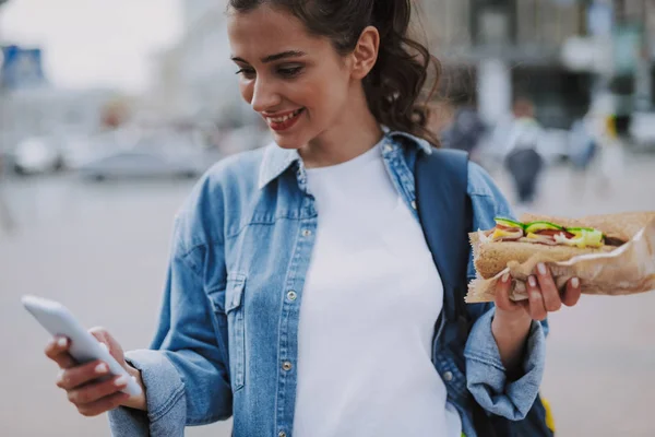 Jolie femelle vérifiant mobile avec hot dog à la main — Photo
