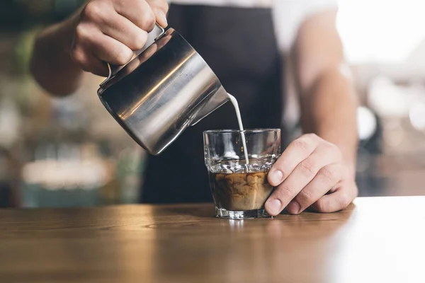 Barista pouring milk from the pitcher into glass of coffee — Stock Photo, Image