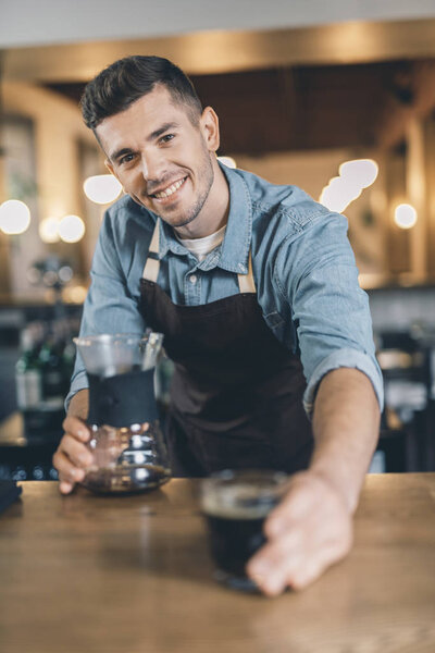 Дружелюбный молодой бариста улыбаясь в камеру и давая залить
