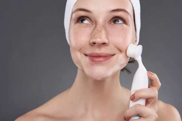 Chica sonriente con pecas usando cepillo de limpieza de poros después del baño —  Fotos de Stock