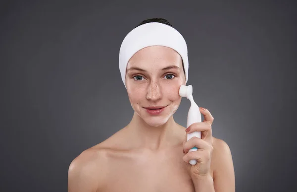 Chica sonriente en diadema blanca usando cepillo de limpieza de poros después del baño —  Fotos de Stock