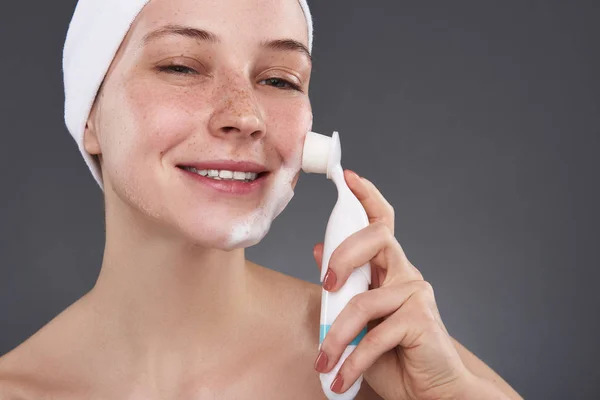 Smiling young woman with freckles using pore cleansing brush after bath