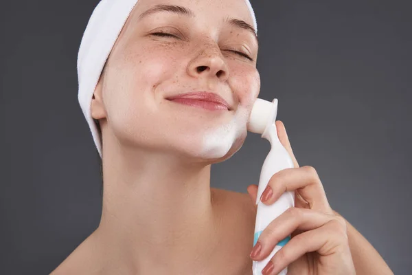Chica sonriente con los ojos cerrados usando cepillo de limpieza de poros después del baño —  Fotos de Stock