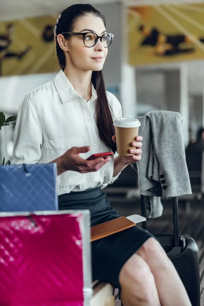Pogodna elegancka kobieta z telefonem pije kawę na lotnisku — Zdjęcie stockowe