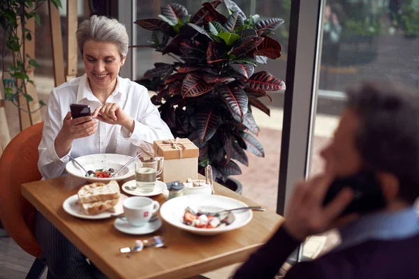 Пожилая улыбающаяся женщина в кафе проверяет смартфон — стоковое фото
