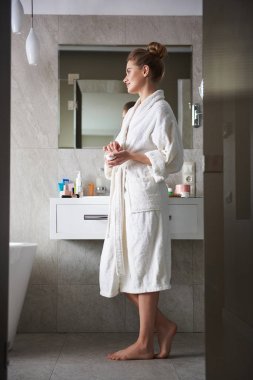 Banyoda elinde yüz kremi tutan gülümseyen genç bayan.