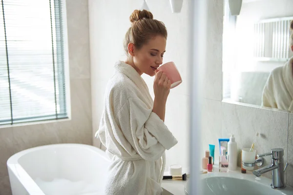 Genç, gülümseyen bayan banyoda kahve içiyor. — Stok fotoğraf