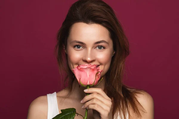 Romantische Frau hält rosa Rose in der Nähe ihres Gesichts und lächelt — Stockfoto