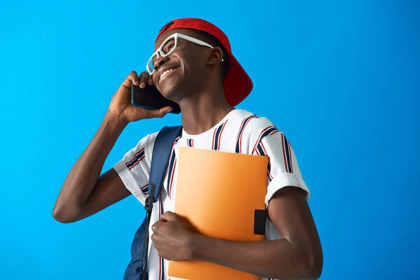 Waist up portrait of Aframerican young student wearing striped t-shirt and talking on smartphone. Isolated on blue background