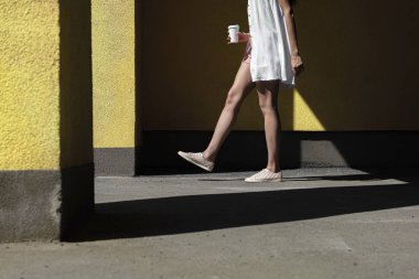 Young woman with take-away drink walking by the yellow wall