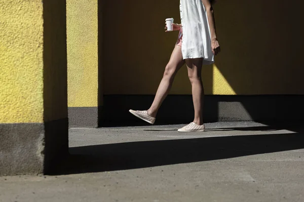 Young woman with take-away drink walking by the yellow wall — Stock Photo, Image