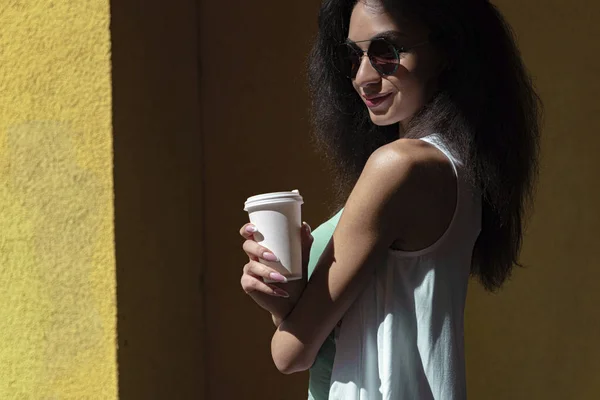 Affascinante giovane donna con tazza di caffè di carta in piedi contro parete gialla — Foto Stock