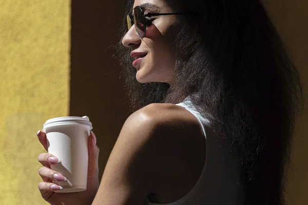 Hermosa mujer joven con taza de café de papel de pie contra la pared amarilla — Foto de Stock