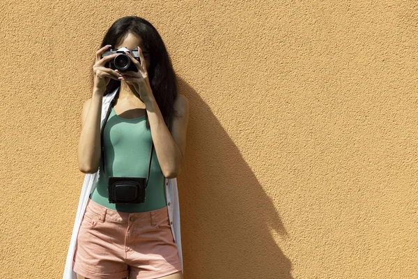 Όμορφη νεαρή γυναίκα με ρετρό κάμερα στέκεται ενάντια ανοιχτό πορτοκαλί τοίχο — Φωτογραφία Αρχείου
