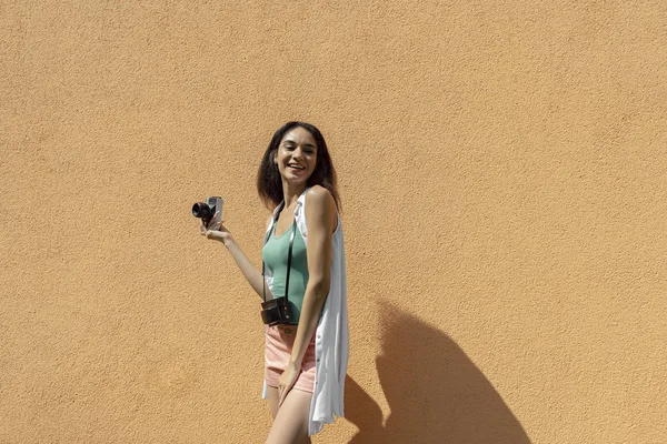Χαρούμενη μελαχρινή κορίτσι με ρετρό κάμερα στέκεται ενάντια φως πορτοκαλί τοίχο — Φωτογραφία Αρχείου
