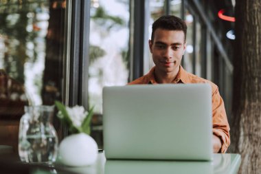 Yakışıklı genç adam masada oturuyor ve dizüstü bilgisayar kullanıyor.