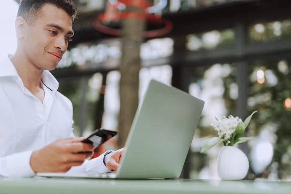 Przystojny młody człowiek z telefon za pomocą laptopa w kawiarni na zewnątrz — Zdjęcie stockowe