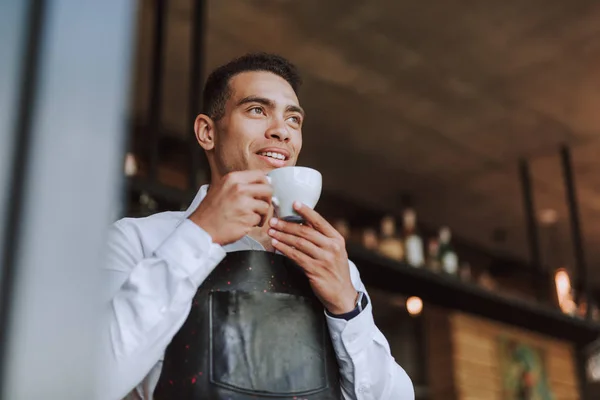 Beau jeune homme en tablier noir buvant du café dans un café — Photo