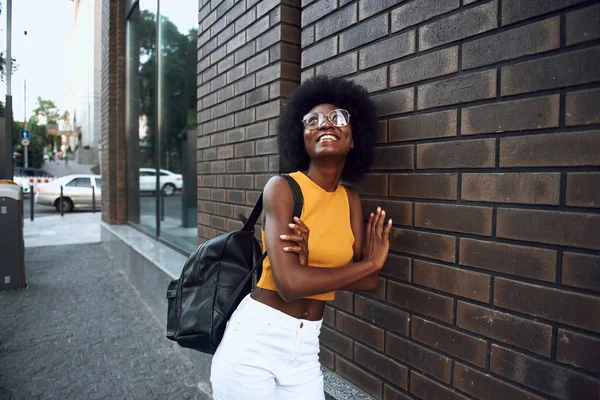 Joyeux jeune femme dans des lunettes appuyé sur le mur de briques dans la ville — Photo