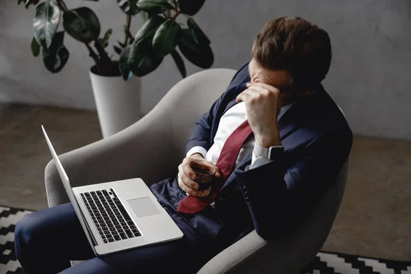 Уставший бизнесмен сидит в кресле с ноутбуком и виски — стоковое фото
