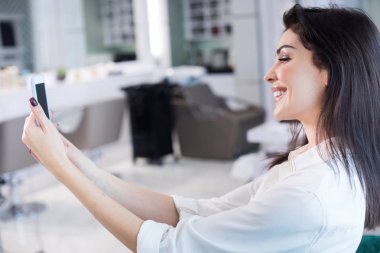 Kuaför salonu kollarında cep telefonu ile gülümseyen bayan Kırpılmış fotoğraf